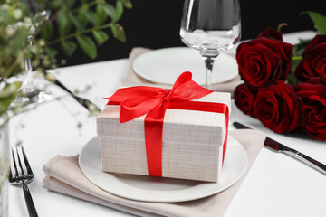 Gift box, plates, cutlery and roses on white table, closeup. Beautiful table setting for romantic...