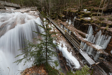 Partly frozen Sabbaday Falls in New Hampshire, a scenic hiking destination with icy cascades, rugged rocks, and a snow-covered wooden walkway. A popular winter trail for nature lovers and adventurers