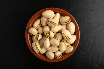 Salted pistachios in a  bowl on a black textured background, isolated, top view, crunchy roasted nuts, tasty and nutritious snack, healthy organic food, high-protein vegetarian ingredient