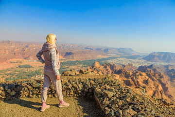 Naklejka premium Blonde tourist pointing at AlUla Old Town from the Harrat Viewpoint, enjoying breathtaking panoramic views of the Saudi Arabian desert landscape in Al-Ula, Madinah province