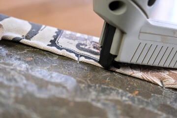 A close-up of a staple gun securing fabric to a hard surface. A staple is partially embedded....