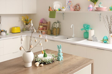 Vase with tree branches, eggs, Easter rabbits and wreath on table in kitchen