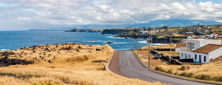 Panoramaaufnahme der Nordk&uuml;ste von S&atilde;o Miguel, Azoren, bei Capelas. Eine schmale Stra&szlig;e f&uuml;hrt sanft hinab zur dramatischen Felsenk&uuml;ste, eingerahmt von goldenem Gras, das im Sonnenlicht leuchtet. 