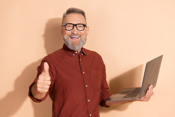 Handsome man showing thumbs up while holding a laptop, wearing an elegant outfit against a beige...