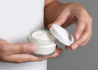 Woman opening cream jar against white shirt and grey background closeup, beauty routine