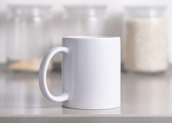 Closeup of white blank coffee mug on grey kitchen table over blured kitchen jars, mockup