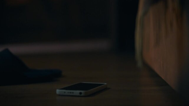 A smartphone lies on the floor close to a sandal in a dimly lit room, suggesting a moment of distraction or forgetfulness. The soft lighting hints at a relaxed evening atmosphere