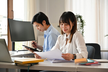 A young woman reviews documents with a smile while her colleague works on a tablet. The organized workspace, reflects efficiency and professional collaboration