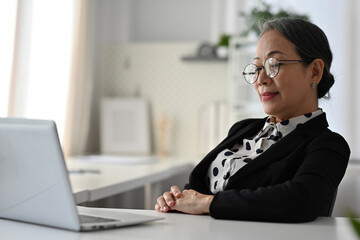 A mature businesswoman in a professional suit leans back with closed eyes, taking a mindful break from work