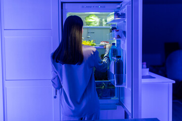 Back view of young woman in sleepwear searching for food in fridge, taking out plate with cake to eat dessert late at night