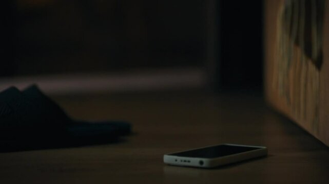 A cell phone lies on the floor next to a pair of slippers. The wooden surface reflects a warm light, suggesting a cozy atmosphere. This action appears to imply forgetfulness or haste