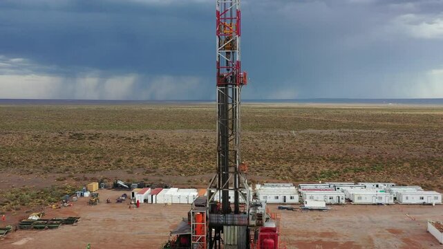Drone performs a backward and upward shot of an oil rig in Vaca Muerta. The sky is stormy and sunny. The rig is red and white. It is in Patagonia, Argentina. Oil and gas extraction location.