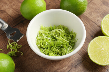 Lime zest in bowl, fresh fruits and zester tool on wooden table, closeup