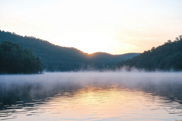 Fototapeta premium Serene misty lake at dawn, surrounded by lush green hills and reflecting the early morning sky.