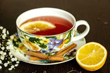 Red  aromatic tea, with a slice of lemon, in a very nicely decorated cup, on a cute saucer, with half a lemon,  small white flowers as decoration