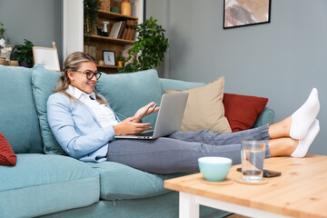 Young beautiful business woman lying on couch using laptop computer in home office apartment,...