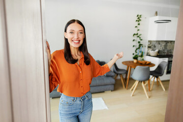 Cheerful European lady opening front door and inviting friends, smiling at camera with welcoming gesture. Real estate concept