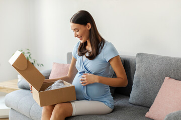Happy young European pregnant woman opening parcel box, receiving baby clothes, looking inside and...