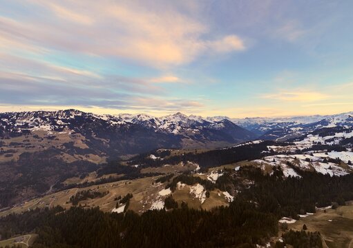 A breathtaking panoramic view of the Alps with snow-capped peaks, rolling green valleys, and a colorful sunset sky. The stunning landscape blends forests, meadows, and mountain villages, creating 