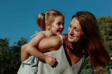 Fototapeta premium A mother carries her young daughter on her back, both smiling brightly under a clear blue sky. They enjoy quality time together in a park filled with greenery