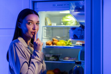 Young woman in sleepwear showing silence gesture standing near open fridge late at night and...