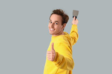 Young man with putty knife showing thumb-up on light background