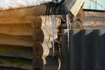 Icicles hang from snow-covered roof of rustic log cabin, creating serene and picturesque winter scene with the sun casting shadows on the wooden structure. .