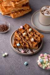 photo Indulge in a delightful serving of fluffy waffles draped in rich chocolate sauce and topped with colorful marshmallows, paired with a frothy cappuccino for a perfect breakfast treat