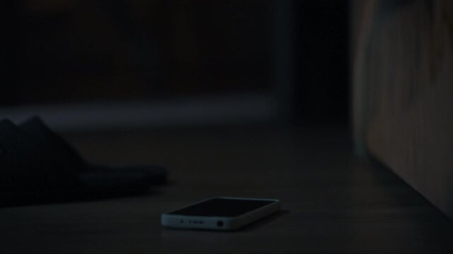 A hand reaches towards a phone that is on the floor in a dimly lit space. The phone screen glows softly, indicating a notification