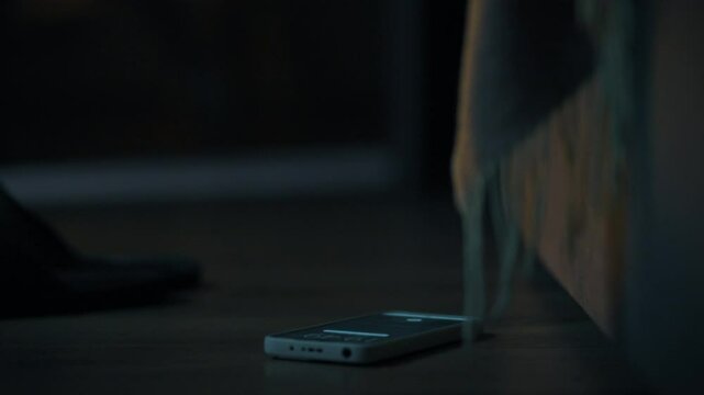 A hand extends towards a smartphone lying on the floor in a dimly lit room. The soft glow from the screen illuminates the surroundings, suggesting a late-night atmosphere