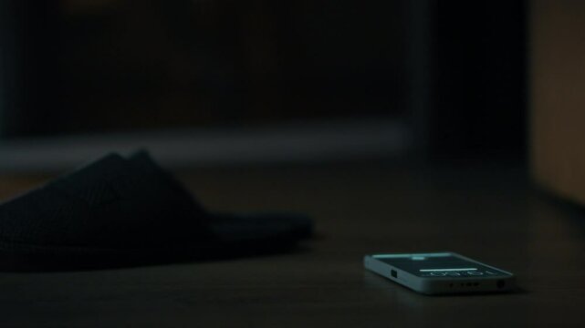 A hand is extended towards a smartphone resting on the floor beside a dark slipper. The dim light creates a calm atmosphere, suggesting a late-night moment of being connected