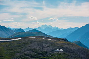 Alpine aerial top view to sunlit big green hill and large rocky mountains silhouettes in bright sun under clouds in blue sky. Scenic layered landscape with high snowy mountain far away in sunny day.