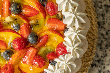 dettaglio macro, da sopra, di una torta di crema e frutta, con pezzi di mirtilli neri, fragole, kiwi e pesche, decorata con crema e panna