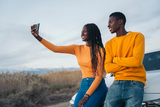 Happy couple taking selfie leaning on car during road trip