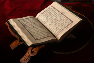 Fototapeta premium An open Quran on a wooden stand against a rich red prayer mat background. The pages display intricate Arabic calligraphy, suitable for themes of spirituality and Islamic culture