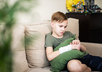 Obraz premium Sad Kid teenager boy at home sitting on sofa with broken finger and suffers from pain. Child hand with a cast.