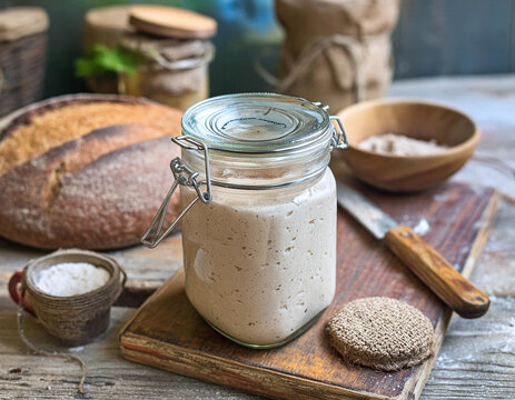 A rustic kitchen scene featuring a glass jar of sourdough starter on a wooden cutting board, surrounded by bread, flour, and kitchen utensils Generative AI