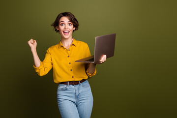 Young happy woman in trendy outfit celebrating success while holding a laptop, against a modern...