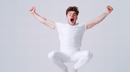 Fototapeta premium Joyful young man in white sportswear jumps and shouts with arms raised against a light gray background.