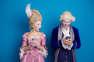 A couple dressed in vintage aristocratic costumes engages with modern technology against a blue...
