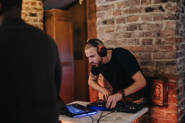 Young man operates DJ equipment, wearing headphones in a warm, rustic bar with a brick wall background. The scene captures artistic expression, creativity, and the ambiance of a nightlife environment.