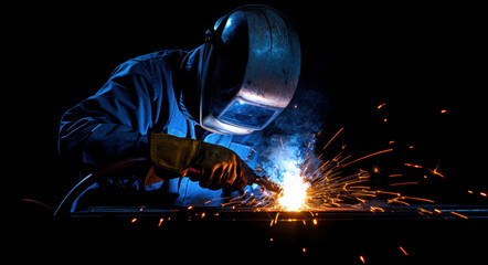 In a dimly lit workshop, a skilled worker focuses on welding with an electric torch, producing bright sparks and soft blue light. The worker wears protective gear for safety while engaged in the task