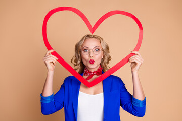 Stylish woman in blue jacket holding red heart shape with cheerful expression against beige...