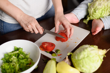 Woman cutting tomato for vegetable salad on kitchen counter, toddler sitting on table nearby holding cabbage, close-up. Concept of vegetarianism, child safety, motherhood, healthy organic food