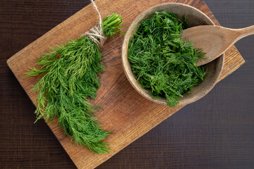 Chopped fresh dill in a wooden plate on a brown kitchen board background next to a bunch of dill. Preparing for freezing. Concept of aromatic herbs for cooking. © pyansetia2008