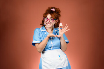 Young smiling waitress, wearing glasses, dress and apron. Holding a cupcake with one hand and pointing to it with the other.