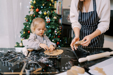 Little girl places cookie cutters on the dough next to her mother kneading the dough