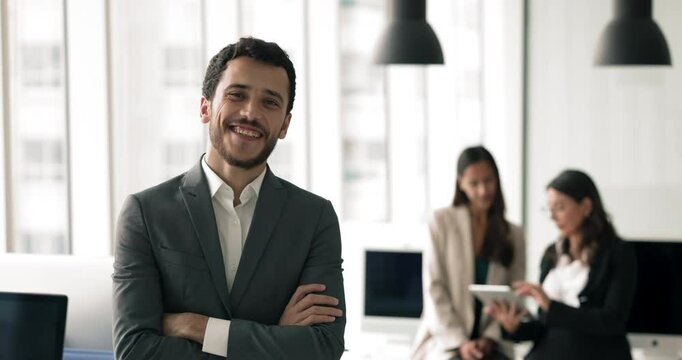 Confident young Arabic businessman posing in open office with colleagues working behind, looking at camera, making hands folded in confidence gesture, getting happy, smiling for portrait