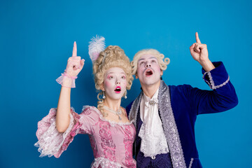 Elegant couple in baroque costumes pointing expressively against a vibrant blue background