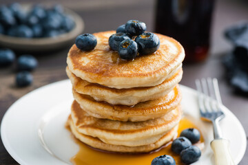 Pancakes with blueberries stack on white ceramic plate served with maple syrup, closeup view, vertical